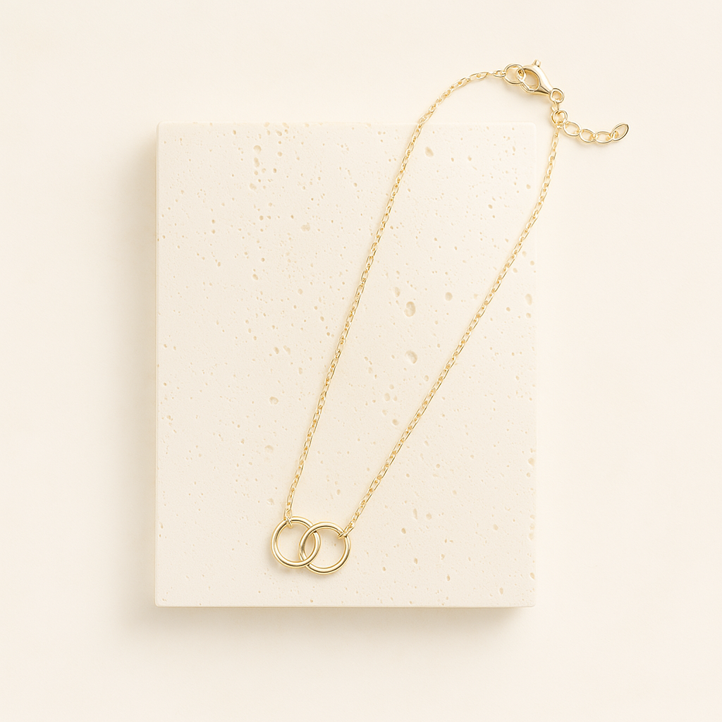 Zarte goldene Halskette mit zwei ineinandergreifenden Ringen, stilvoll arrangiert auf einer hellen Travertinplatte vor cremefarbenem Hintergrund – minimalistisches und elegantes Produktfoto im Quadratformat.