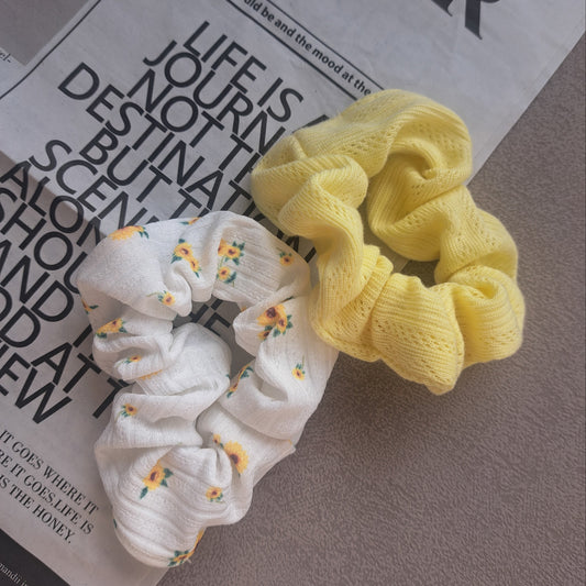 Zwei stilvolle Scrunchies – eines sonnengelb und eines weiß mit kleinen Sonnenblumen – liegen auf einem weißen Untergrund mit dekorativer Zeitung, elegant und hell inszeniert.