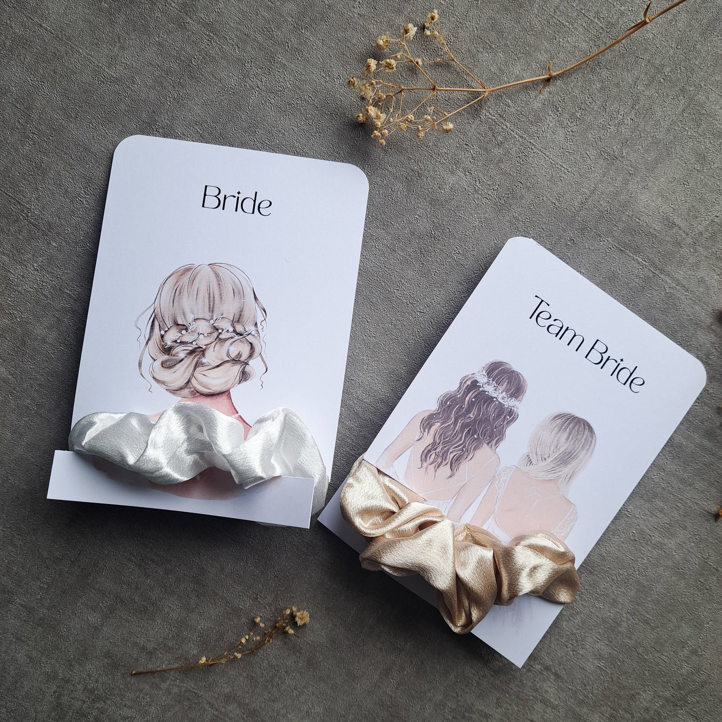 Zwei elegante Haargummis aus Satin auf illustrierten Karten mit der Aufschrift „Bride“ und „Team Bride“, dekoriert mit getrockneten Blüten auf hellem Untergrund.