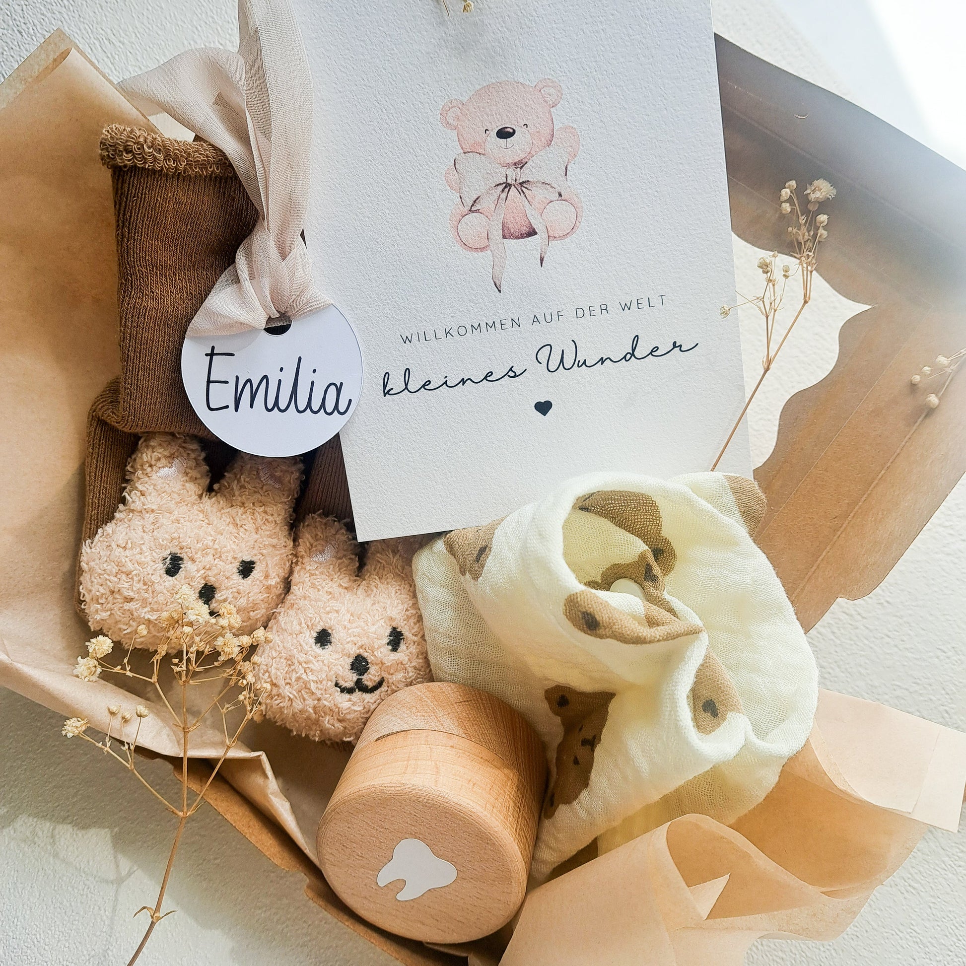 Geschenkset zur Geburt mit personalisiertem Namenslabel „Emilia“, Musselin-Halstuch mit Bärenmotiv, Babysöckchen, Zahndose und Karte „Willkommen auf der Welt, kleines Wunder“ – liebevoll arrangiert auf hellem Untergrund