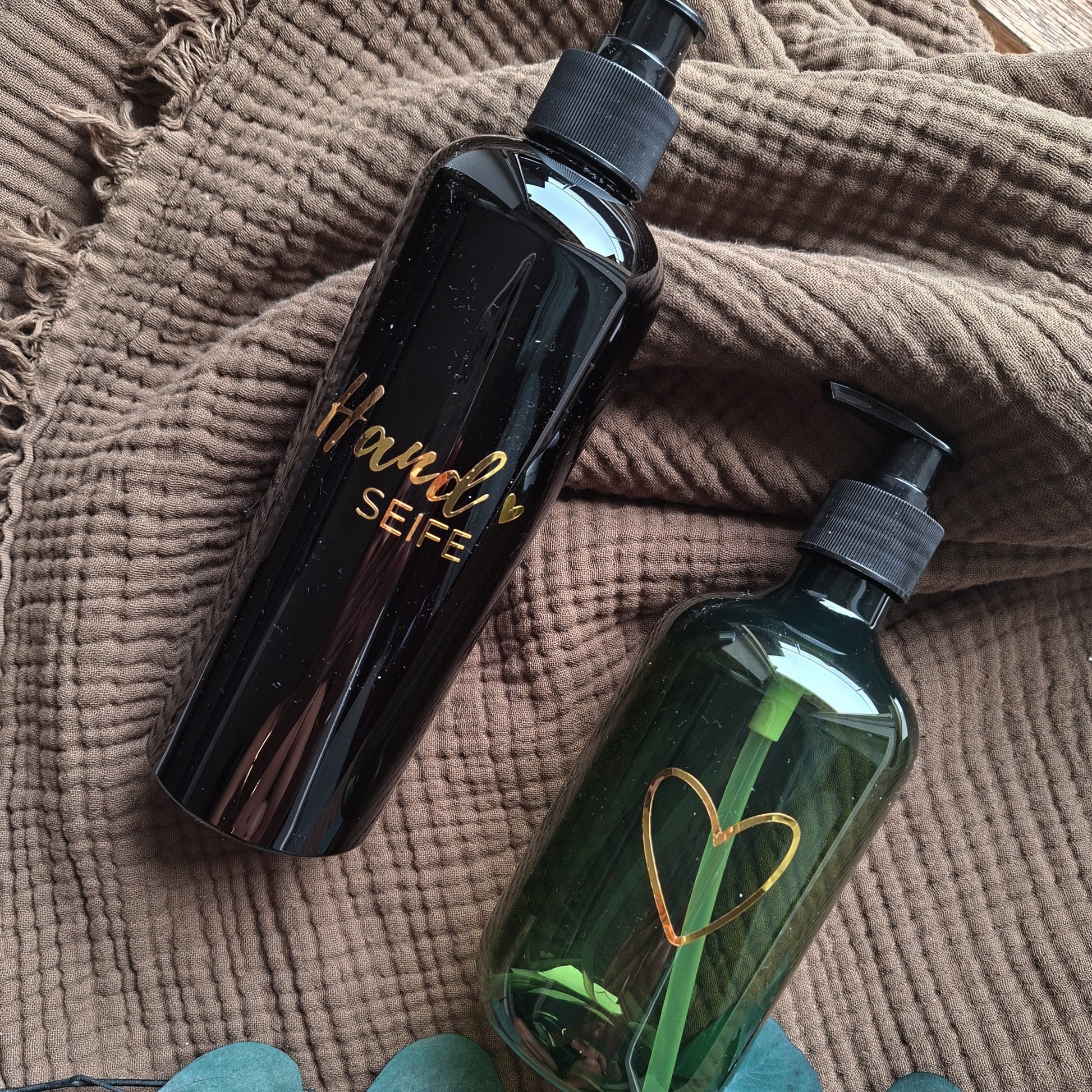 Zwei stilvolle Seifenspender auf braunem Handtuch – schwarzer Spender mit goldener „Handseife“-Beschriftung und grüner Spender mit goldenem Herzsymbol, dekoriert mit Eukalyptusblättern – modernes Badezimmer-Accessoire