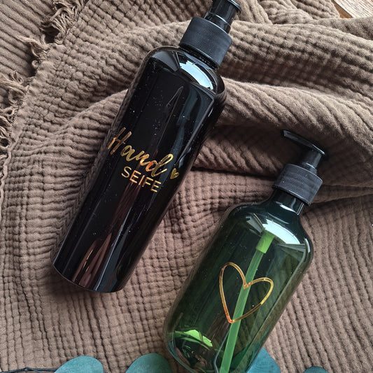 Zwei stilvolle Seifenspender auf braunem Handtuch – schwarzer Spender mit goldener „Handseife“-Beschriftung und grüner Spender mit goldenem Herzsymbol, dekoriert mit Eukalyptusblättern – modernes Badezimmer-Accessoire